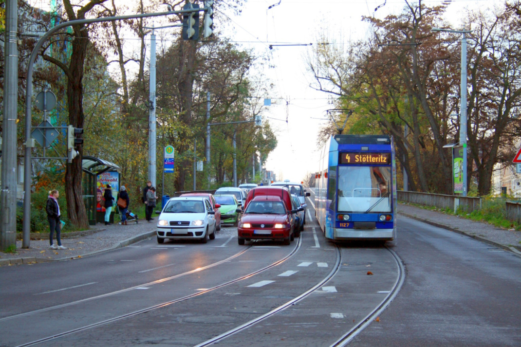 Straßenbahn und wartende PKW auf Straße, Haltestelle mit Passanten.