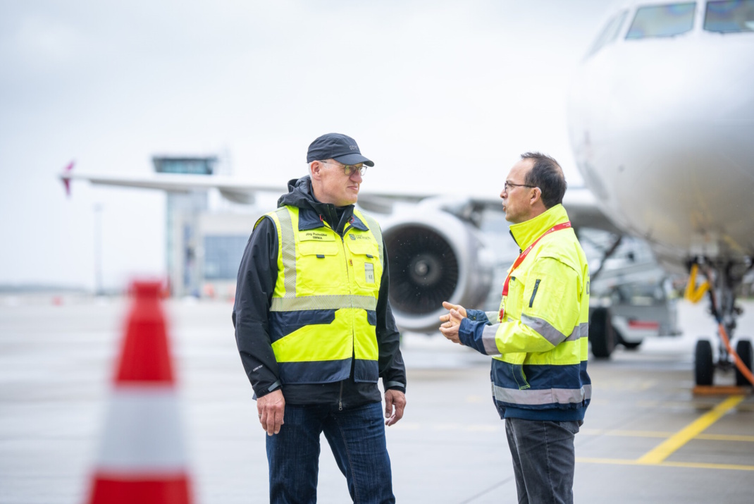 Zwei Männer in grellen Arbeitsjacken sprechen miteinander, im Hintergrund ist ein stehendes Flugzeug erkennbar.