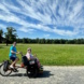 Personen mit Rikscha vor grüner Wiese, Wolken und blauer Himmel.