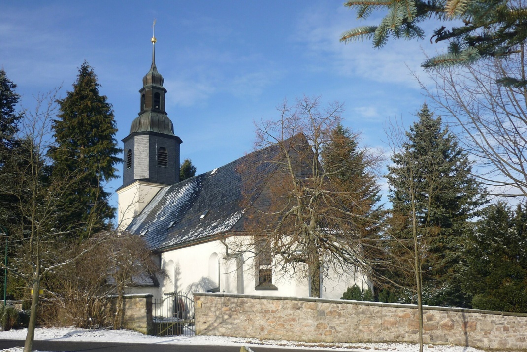 Weiße Kirche mit dunklem Dach, umgeben von Mauer, Bäumen und blauem Himmel.