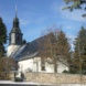 Weiße Kirche mit dunklem Dach, umgeben von Mauer, Bäumen und blauem Himmel.