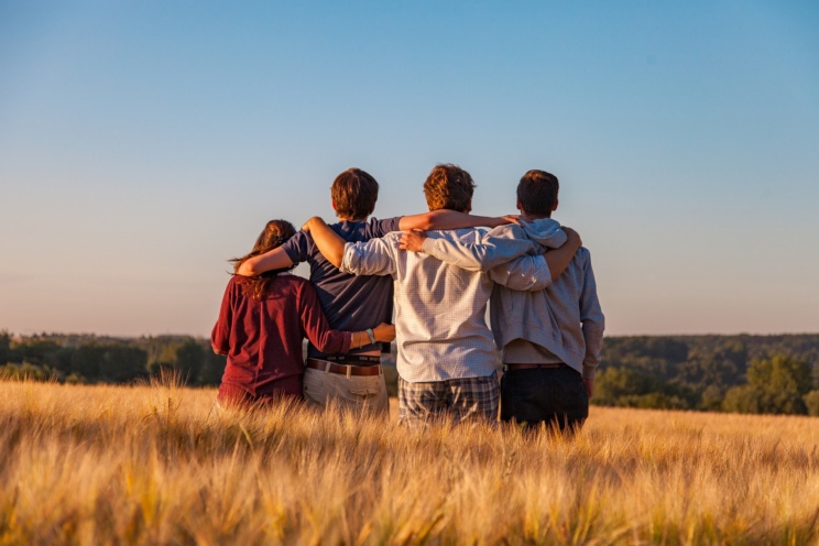 Vier junge Menschen sind mit Rückenansicht zu sehen, wie sie Arm in Arm auf einem Feld stehen.