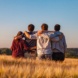 Vier junge Menschen sind mit Rückenansicht zu sehen, wie sie Arm in Arm auf einem Feld stehen.