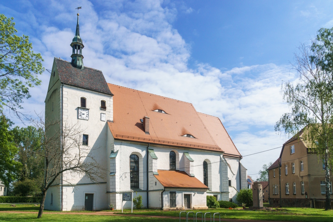 Helle Kirche mit rotem Dach und umgeben von Bäumen, blauer Himmel.