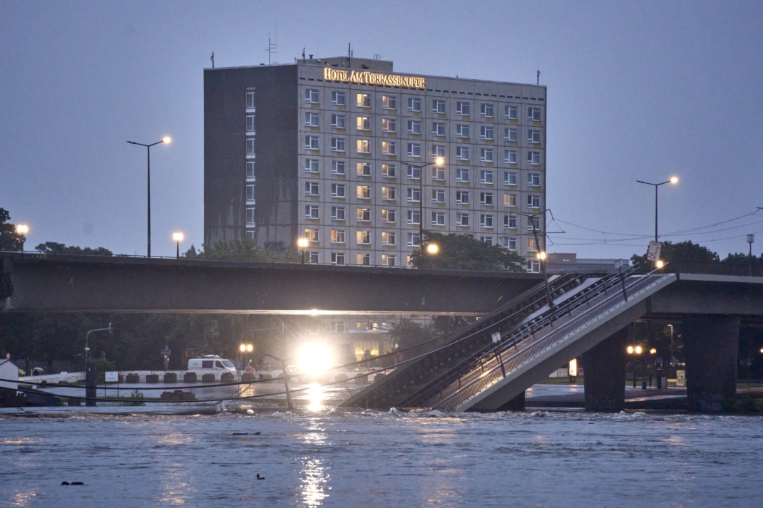 Eingestürzter Teil einer Brücke in der Dämmerung, die über einen Fluss führt, beleuchtetes Gebäude und Scheinwerfer.