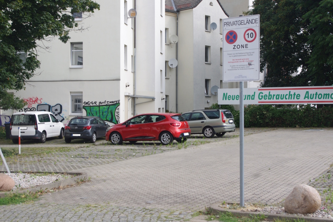 Zu sehen ist ein Parkplatz mit abgestellten PKW vor einer Hauswand, Aufnahme aus größerer Distanz.