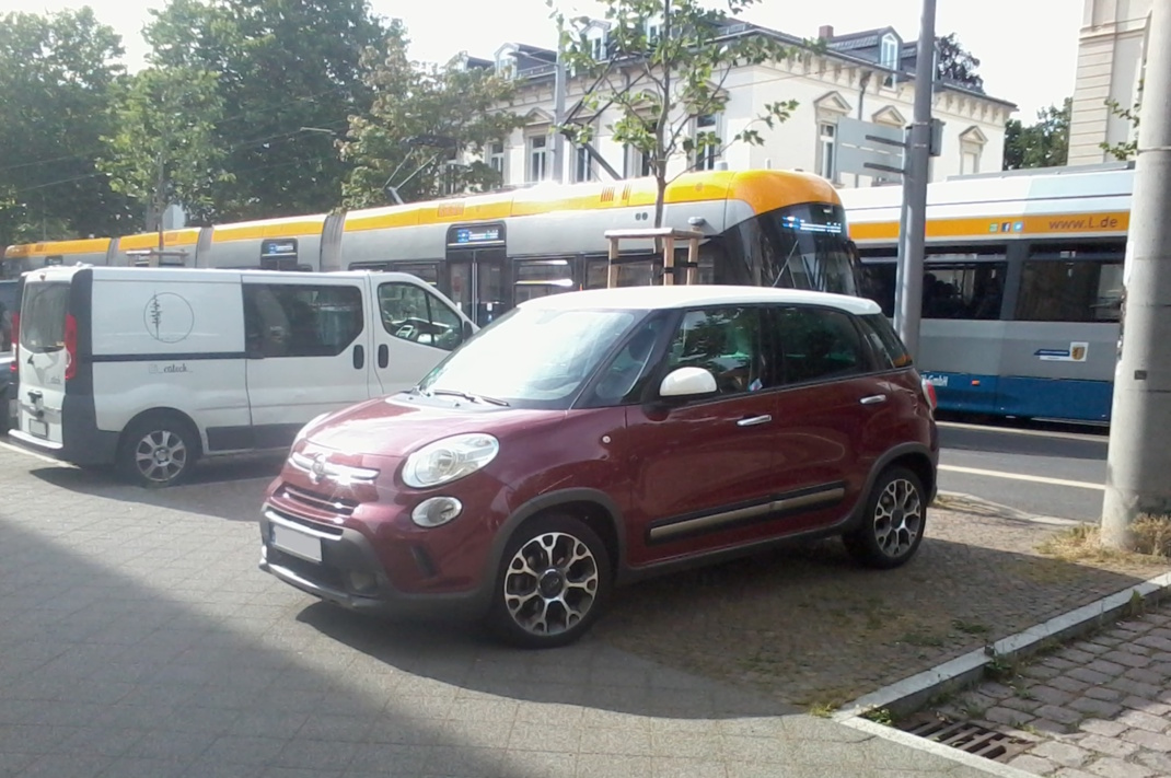 Parkende Autos stehen in der Sonne auf dem Gehweg, dahinter rollen Straßenbahnen durch eine Hauptstraße.