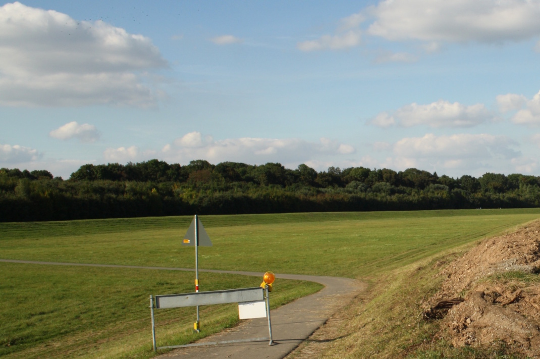 Landschaft mit Wiese, Erdhügel, asphaltiertem Weg, Wolken und blauem Himmel.