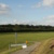 Landschaft mit Wiese, Erdhügel, asphaltiertem Weg, Wolken und blauem Himmel.