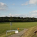 Landschaft mit Wiese, Erdhügel, asphaltiertem Weg, Wolken und blauem Himmel.