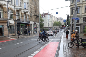 Städtische Haltestelle mit rot markiertem Radweg auf der Hauptstraße, Radlern und Autofahrern.