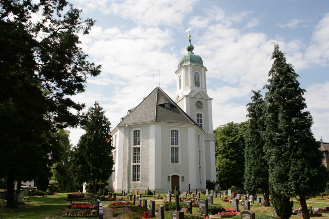 Friedhof, weißes Kirchenhaus, Bäume und blauer Himmel mit Wolken.