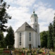 Friedhof, weißes Kirchenhaus, Bäume und blauer Himmel mit Wolken.