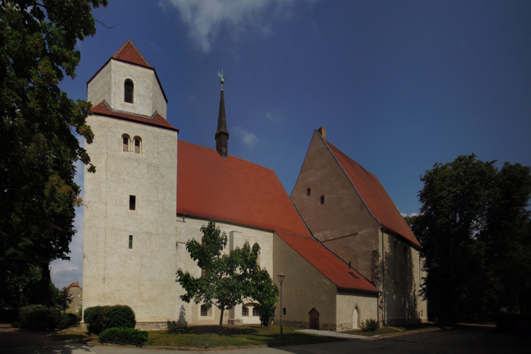 Helles Kirchengebäude mit roten Dächern, Bäumen und blauem Himmel.