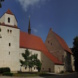 Helles Kirchengebäude mit roten Dächern, Bäumen und blauem Himmel.