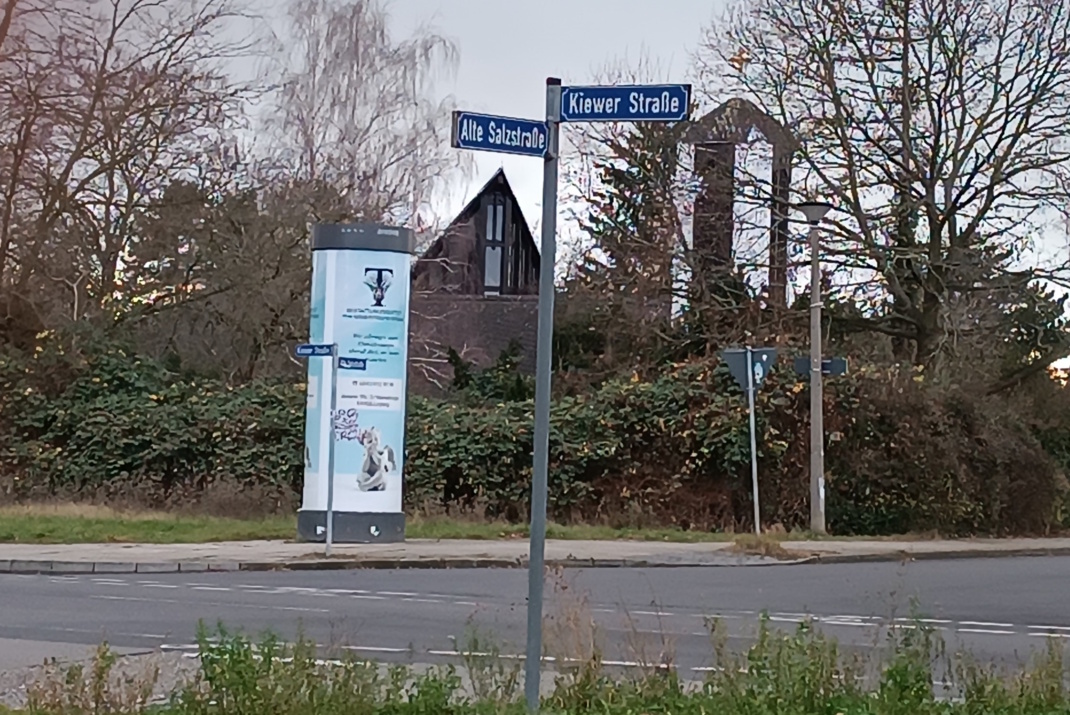 Straßenschilder an einer Ecke, zu sehen sind auch eine Litfaßsäule und Gebüsch.