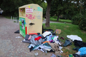 Öffentlicher Kleidercontainer, neben dem zahlreiche Textilien aufgehäuft liegen