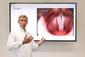 Prof. Michael Fuchs demonstriert die Funktion der Stimmlippen beim Singen vor einem Bild.